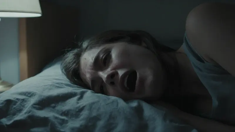 Mujer con la boca abierta en un grito silencioso, en una habitación oscura con sábanas revueltas y luz tenue de una lámpara de noche.
