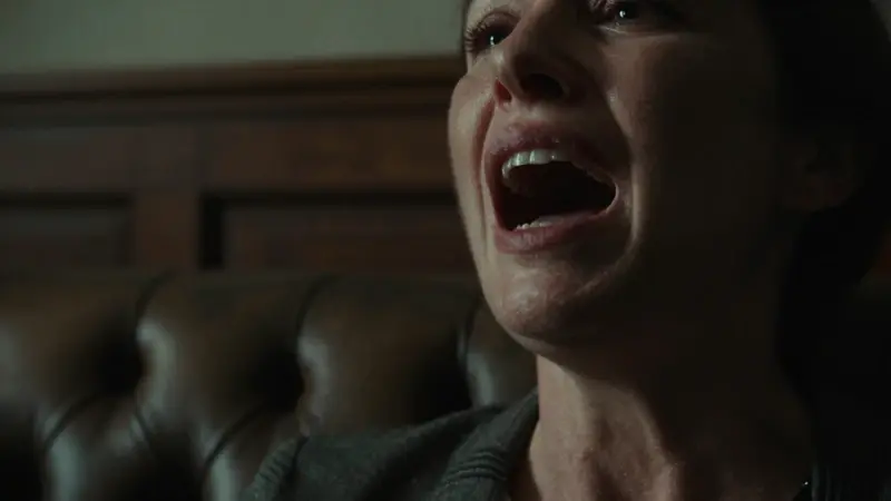 Primer plano de una mujer con expresión de angustia, intentando gritar en un consultorio psicoanalítico tenuemente iluminado, con un diván de cuero y paneles de madera oscura.
