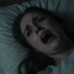 Primer plano de una mujer con expresión de angustia, intentando gritar en la oscuridad de su dormitorio. Las sábanas están revueltas y la iluminación tenue sugiere un ambiente opresivo y de pesadilla.