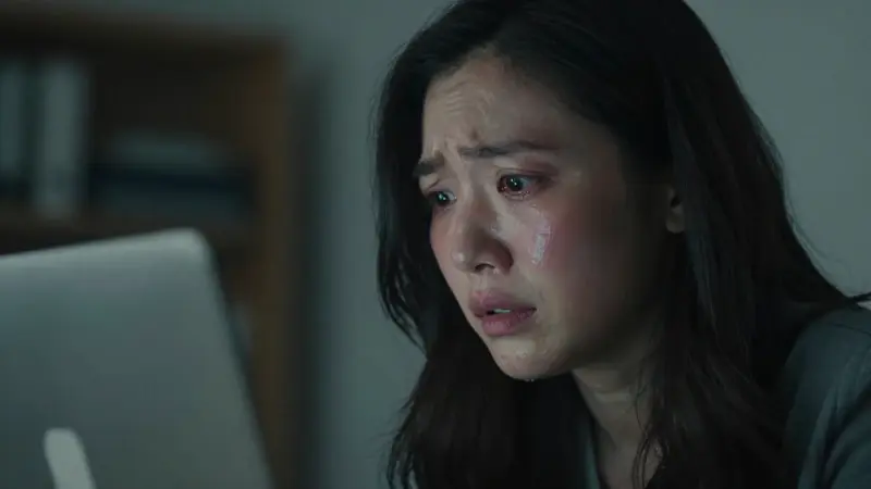 Primer plano de una mujer sonrojada y con expresión de ansiedad frente a un portátil en una oficina tenuemente iluminada por la noche.