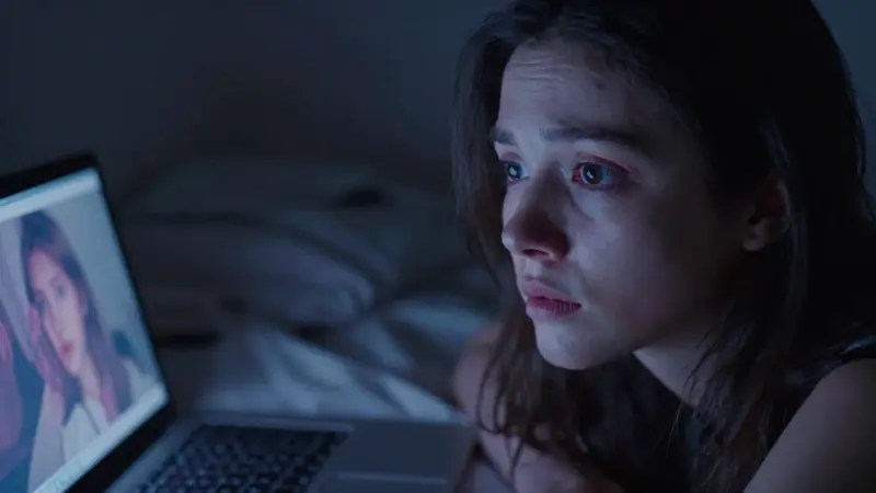 Joven mujer despierta en la noche, con expresión de miedo y la luz de un portátil proyectando imágenes oníricas distorsionadas en su rostro.