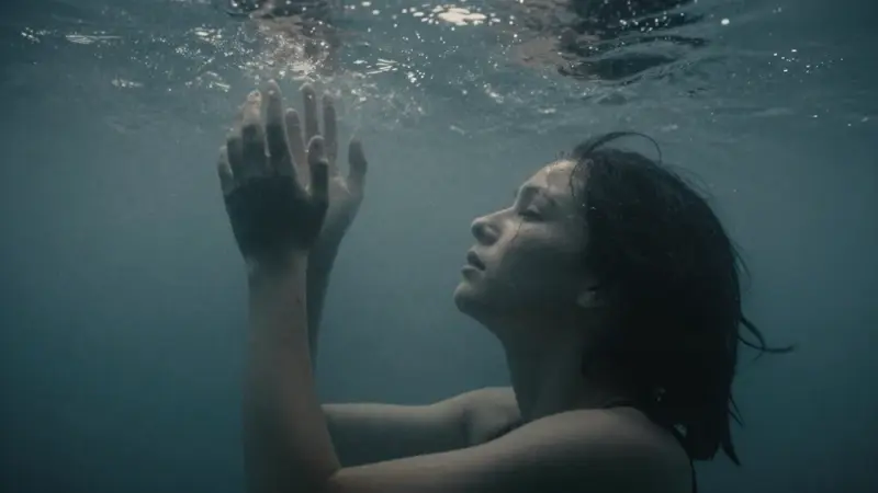 Figura sumergida silueteada contra una luz difusa bajo el agua, con expresión de calma y desesperación, y manos extendidas hacia arriba en un entorno turbio y azulado.
