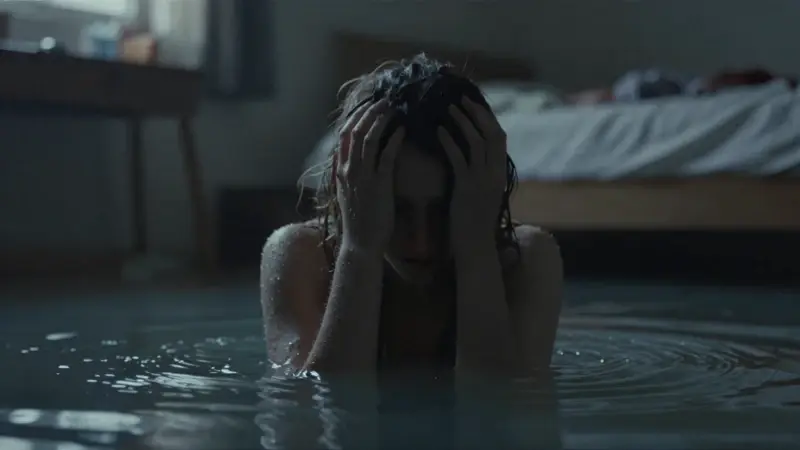Silueta sumergida en agua turbia con un interior de dormitorio difuminado al fondo, evocando una atmósfera de ansiedad y opresión.