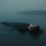 Persona hundiéndose lentamente en aguas oscuras y turbias, con una costa borrosa a lo lejos. Ambiente melancólico y sensación de aislamiento.