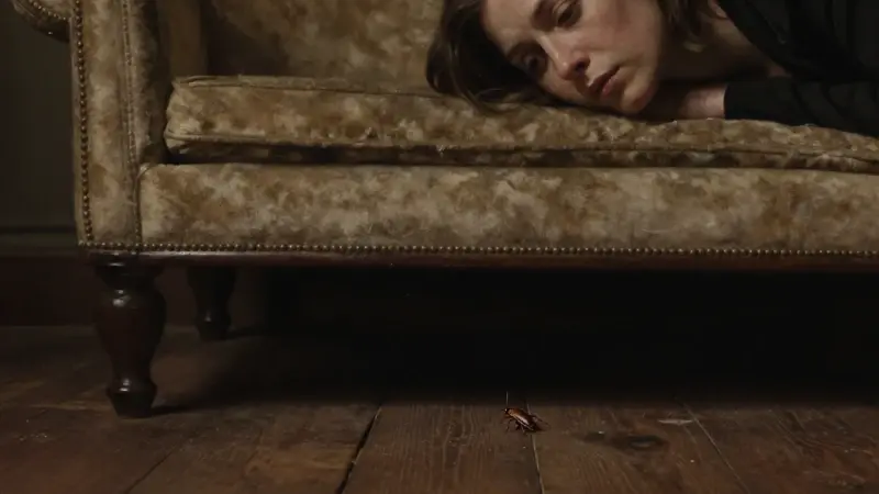 Primer plano de una mujer pensativa, con un diván de psicoanálisis antiguo en segundo plano. Una cucaracha camina sobre una tabla de madera cercana, en una atmósfera melancólica y con iluminación dramática.