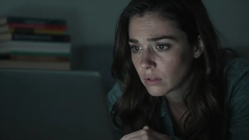 Mujer preocupada mirando una laptop con una pantalla iluminada en un dormitorio oscuro, posiblemente buscando información sobre sueños.