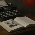 Estudio tenuemente iluminado con un retrato de Sigmund Freud en un libro abierto, rodeado de páginas de diario de sueños y una máquina de escribir antigua. Un globo rojo, alusivo a la novela "It" de Stephen King, se vislumbra en el fondo.