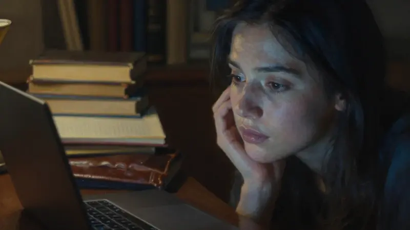 Mujer mirando la pantalla de un portátil en un estudio oscuro, rodeada de libros y diarios de sueños, posiblemente investigando el significado de soñar con Blancanieves.