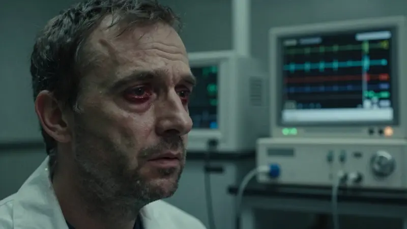 Primer plano de un hombre de mediana edad con rostro cansado y ojos enrojecidos, rodeado de equipos de monitoreo en un laboratorio. Visualización de patrones de ondas cerebrales en un electroencefalograma.