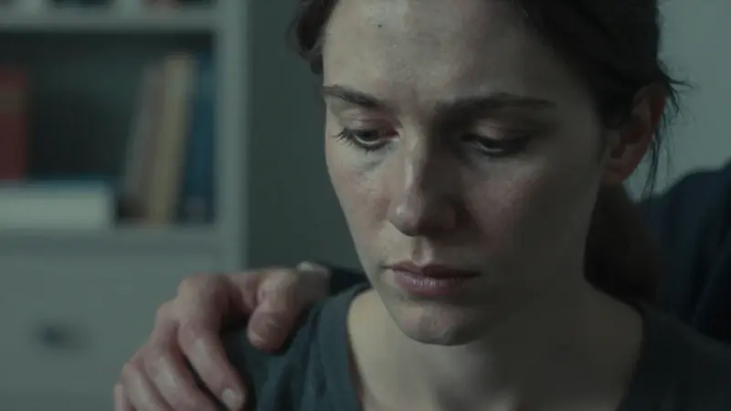 Mujer con mirada introspectiva en un consultorio de terapia, recibiendo un gesto de apoyo en el hombro. Fondo difuminado con estanterías.