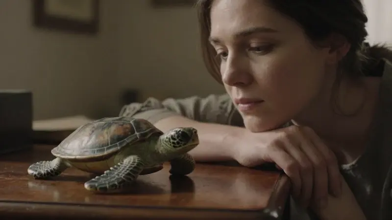 Mujer pensativa mirando una escultura realista de tortuga marina sobre un escritorio de madera antigua en una habitación con luz tenue.
