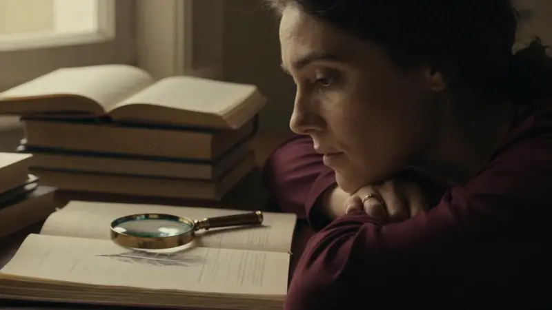 Mujer pensativa en un estudio con libros sobre análisis de sueños, una lupa sobre dibujos de plumas y luz cálida entrando por la ventana.