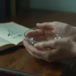 Manos sosteniendo agua clara sobre un escritorio antiguo, con un diario abierto y bocetos borrosos en el fondo. Ambiente introspectivo y cálido.