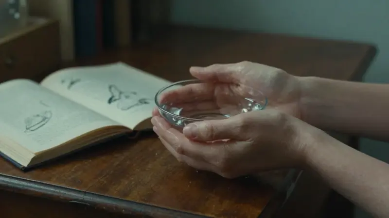 Manos sosteniendo agua clara sobre un escritorio antiguo, con un diario abierto y bocetos borrosos en el fondo. Ambiente introspectivo y cálido.