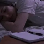Primer plano de una mujer con expresión preocupada, a medio ocultar en la penumbra de un dormitorio desordenado con una libreta de sueños y un bolígrafo sobre la mesita de noche.