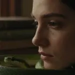 Primer plano de rostro femenino parcialmente cubierto por una serpiente verde esmeralda, en un estudio antiguo con libros apilados y luz tenue.