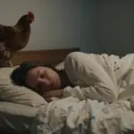 Mujer durmiendo en una habitación tenue con una gallina marrón posada en el pie de la cama.