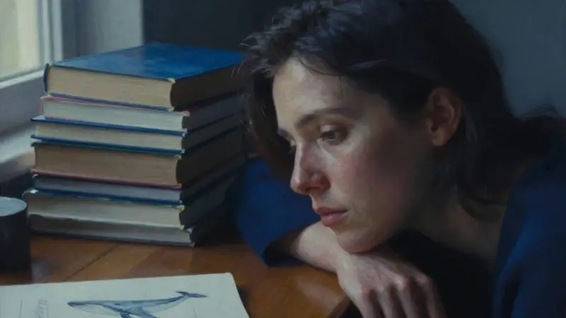 Mujer pensativa en un estudio con libros sobre análisis de sueños, con un cuaderno mostrando bocetos de ballenas. Predominan los tonos azules y morados, creando una atmósfera melancólica y serena.