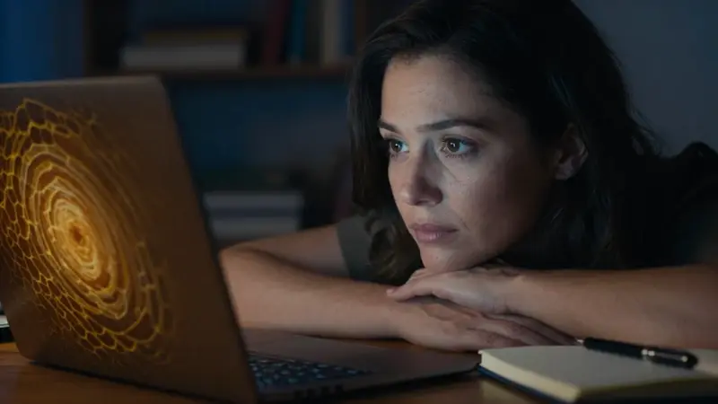 Mujer pensativa frente a un portátil con patrones que recuerdan al panal, en un estudio tenuemente iluminado, posiblemente analizando el significado de sus sueños.