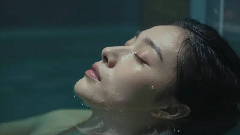 Mujer con rostro sereno, parcialmente sumergida en agua oscura y translúcida, con los ojos cerrados en un estado onírico, y un acuario difuminado de fondo.