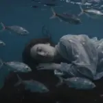 Mujer dormida rodeada de bancos de peces plateados en un ambiente submarino azul oscuro y onírico.