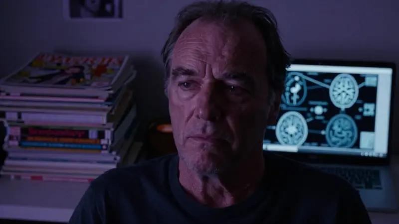 Retrato de un hombre cansado, parecido a Jack Nicholson, rodeado de cómics de Sleepwalker y una computadora que muestra imágenes de sueños y escaneos cerebrales en un escritorio desordenado.