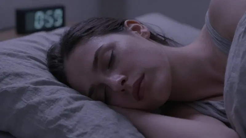 Mujer durmiendo plácidamente en una cama arrugada, con una luz tenue iluminando su rostro y destacando una sutil tensión muscular. El ambiente es tranquilo e introspectivo, con un reloj digital borroso en el fondo.