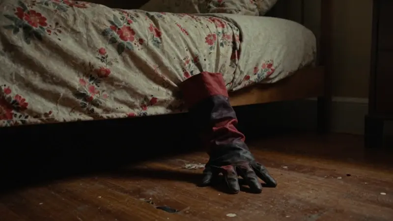 Guante con garras de Freddy Krueger asomando por debajo de una cama en un dormitorio oscuro y desolado, evocando una atmósfera amenazante y nostálgica de los años 80.