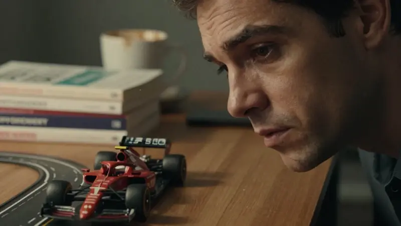 Hombre observando un circuito de Fórmula 1 en miniatura sobre un escritorio lleno de libros de psicología y una taza de café.