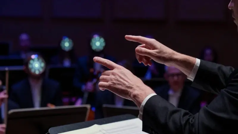 Manos de un director de orquesta guiando un conjunto de relojes biológicos luminosos en lugar de instrumentos musicales, en un ambiente sofisticado y dramáticamente iluminado.