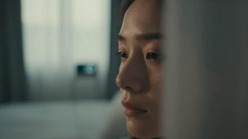 Mujer contemplativa en una habitación a primera hora de la mañana, con un reloj digital borroso al fondo y luz tenue filtrándose por las cortinas.