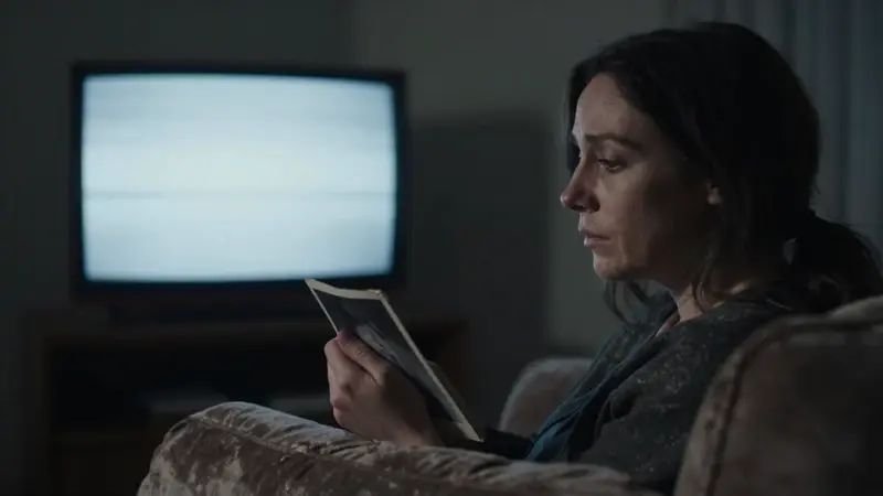 Mujer angustiada abrazando una foto antigua en un salón oscuro, con la televisión encendida mostrando estática.