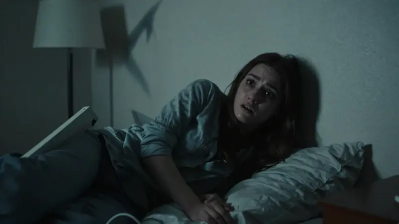 Joven angustiada en un dormitorio oscuro y desordenado, con una sombra amenazante en la pared.