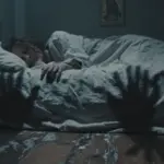 Persona durmiendo inquietamente en un dormitorio tenuemente iluminado, rodeada de sombras que recuerdan a manos que intentan alcanzarla. Iconografía religiosa antigua se vislumbra sutilmente en el fondo.