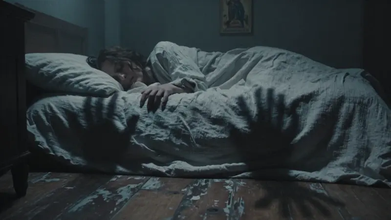 Persona durmiendo inquietamente en un dormitorio tenuemente iluminado, rodeada de sombras que recuerdan a manos que intentan alcanzarla. Iconografía religiosa antigua se vislumbra sutilmente en el fondo.