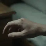 Mano humana con una garrapata incrustada en la piel, en un ambiente tenue de dormitorio con noche y mesita de madera desgastada.