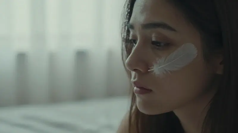 Mujer pensativa en su dormitorio, con una pluma blanca sobre la mejilla y luz tenue de la mañana, evocando un ambiente de introspección.