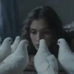 Primer plano de una mujer con el rostro parcialmente cubierto por una bandada de palomas blancas en un dormitorio tenuemente iluminado.