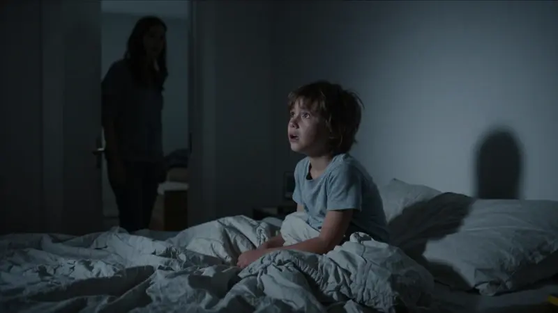 Niño asustado sentado en la cama en medio de la noche, con un progenitor preocupado en el marco de la puerta.
