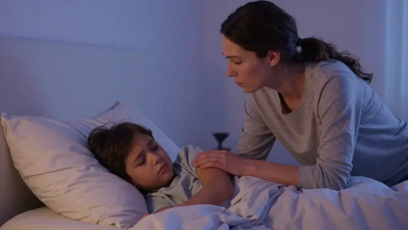 Madre preocupada consuela a su hijo mientras duerme, visiblemente angustiado por un terror nocturno en un dormitorio tenuemente iluminado.