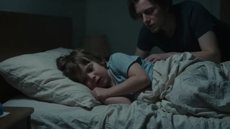 Niño pequeño retorciéndose en la cama durante un terror nocturno, con sus padres preocupados observando a su lado en un dormitorio tenuemente iluminado.