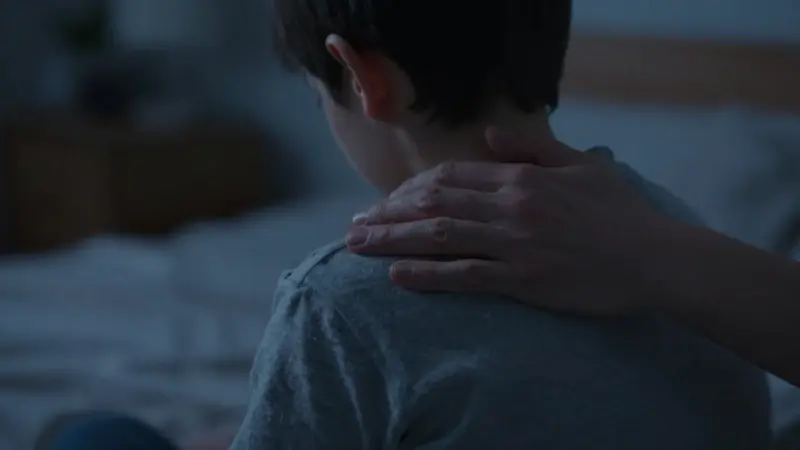 Una mano de adulto acaricia suavemente el hombro de un niño en un dormitorio oscuro, transmitiendo calma y protección.