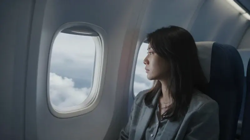 Interior de avión con una mujer mirando pensativa por la ventana un cielo nublado.