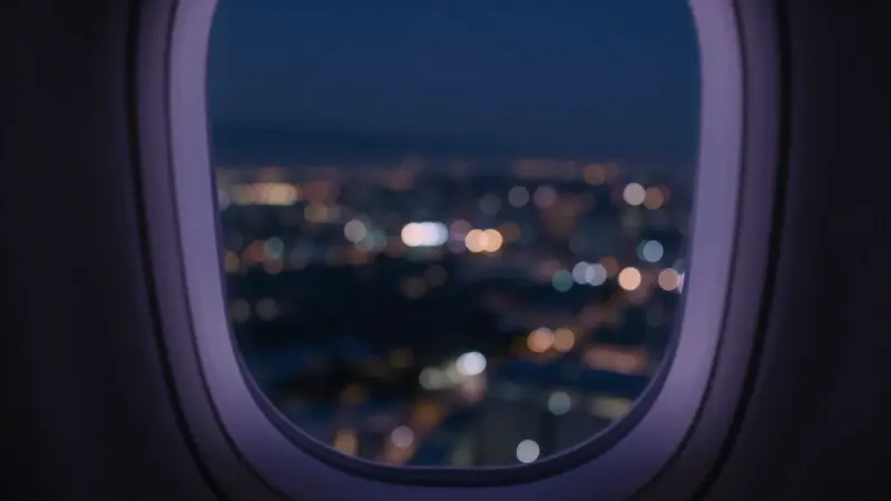 Vista desde la ventana de un avión a la noche, con luces borrosas de la ciudad abajo y tonos azules y morados melancólicos.