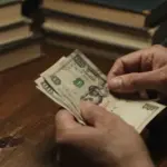 Manos angustiadas contando billetes gastados sobre un escritorio antiguo, con libros borrosos al fondo. Ambiente melancólico y tenue.