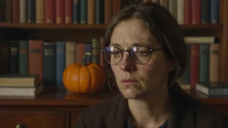 Mujer preocupada en estudio con estanterías llenas, con un reflejo sutil de una calabaza en sus gafas, atmósfera melancólica y tonos otoñales.