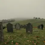 Cementerio antiguo bajo un cielo nublado, con lápidas desgastadas y colinas verdes. Una figura solitaria contempla el paisaje otoñal entre la niebla.