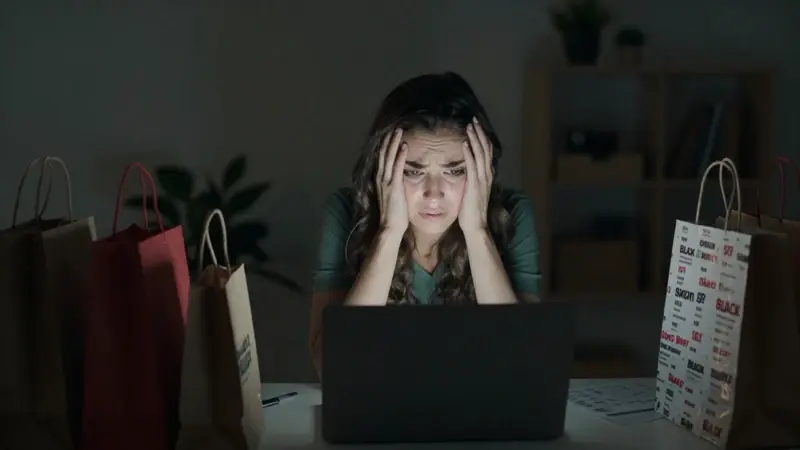 Mujer frustrada frente a su laptop en la noche, rodeada de bolsas de compras, mientras navega por ofertas caóticas del Black Friday.