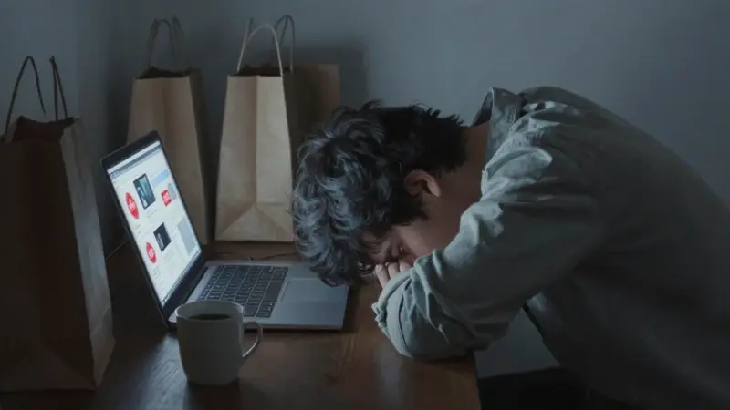 Persona abrumada frente a un escritorio lleno de bolsas de compras y un portátil con anuncios de ofertas, en una habitación tenuemente iluminada.