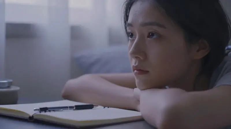 Retrato de una mujer pensativa en su dormitorio, con un cuaderno y bolígrafo sobre la mesita de noche, evocando la reflexión sobre los sueños y la memoria.